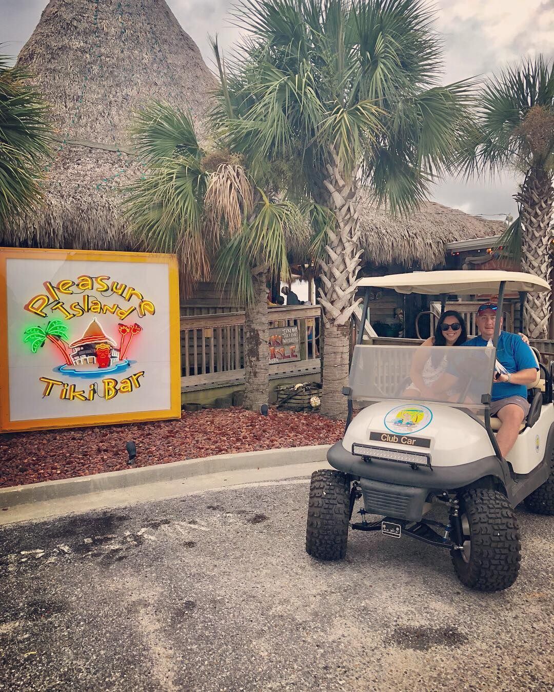 A man and woman sitting in a golf cart parked in front of a thatched-roof tiki bar with a colorful sign.