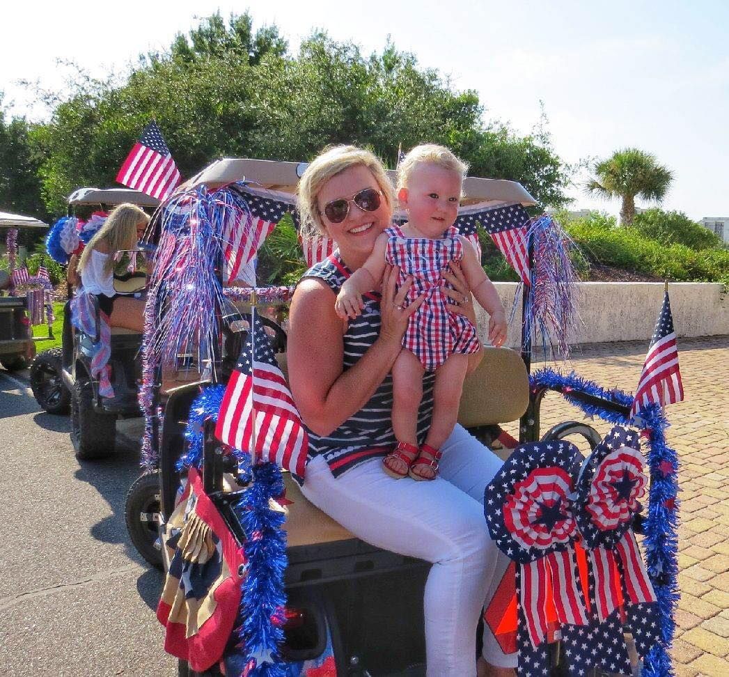 A person holds a child in a festive, USA-themed decorated golf cart at an outdoor sunny parade.