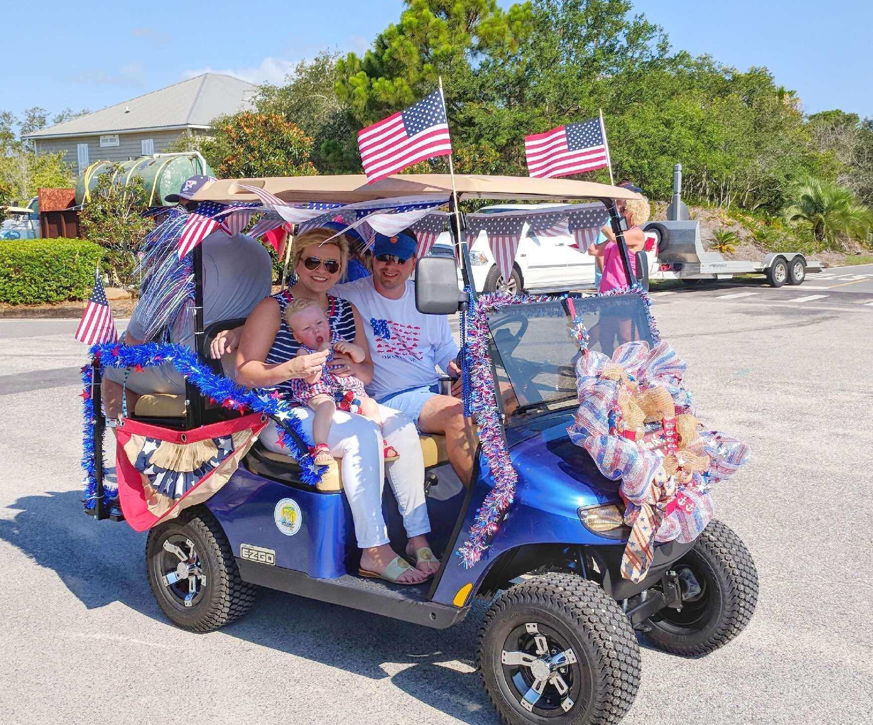A family poses on a decorated blue golf cart adorned with American flags, bunting, and festive bows in a parking lot.