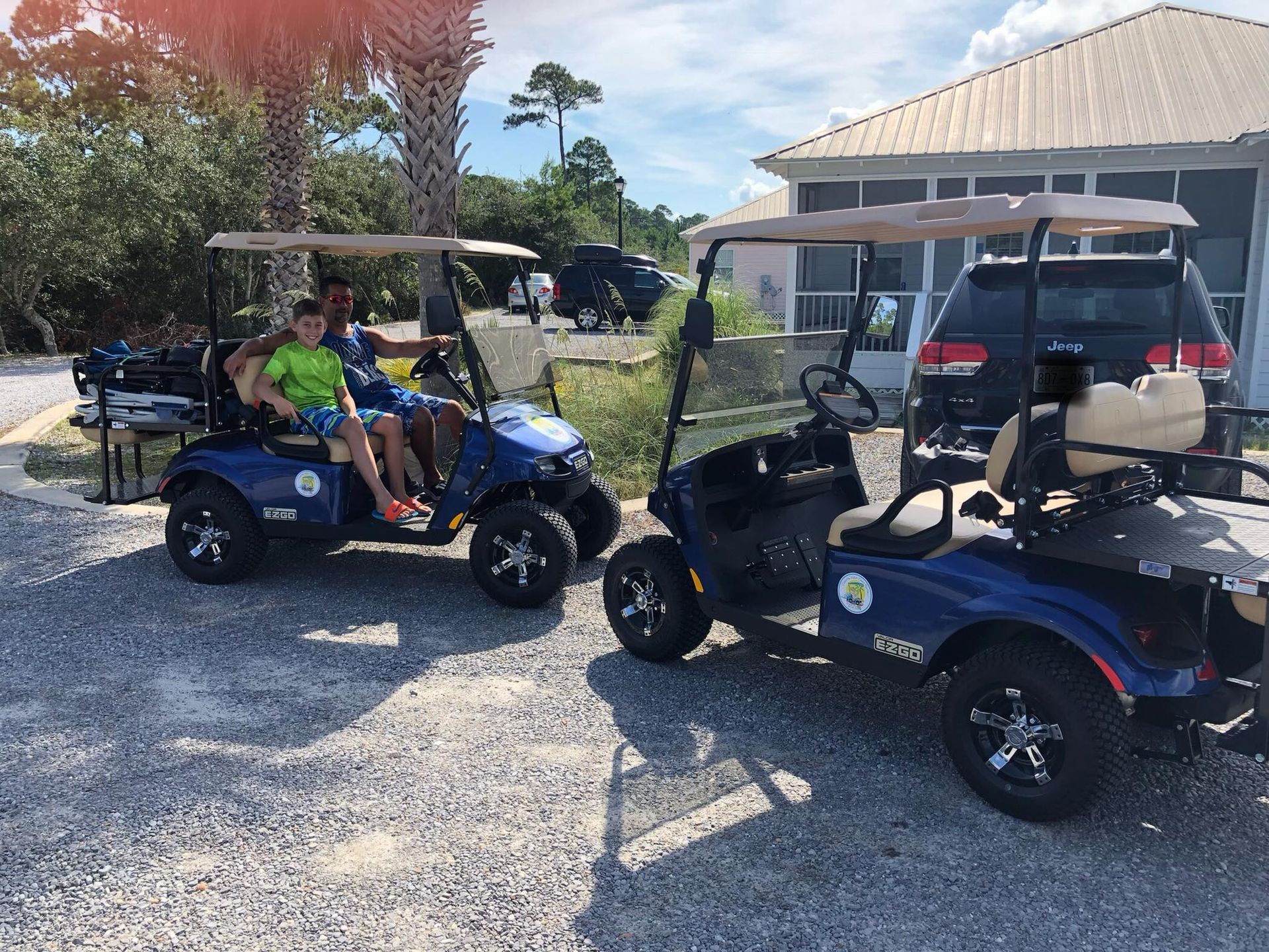 Two blue golf carts parked on gravel near a house, with two people seated in the left cart.