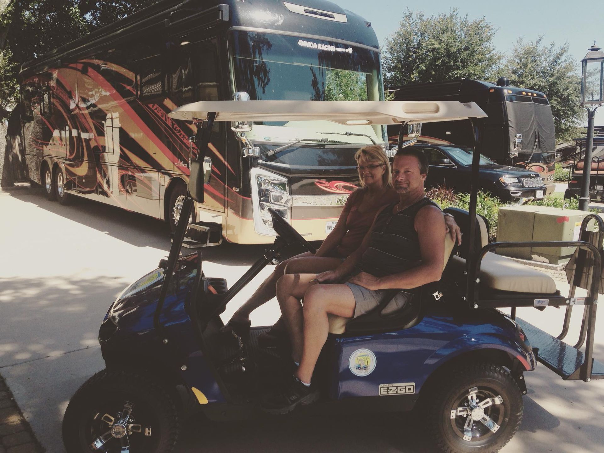 A couple sits on a blue golf cart at a sunny RV park, with a large, patterned motorcoach parked in the background.