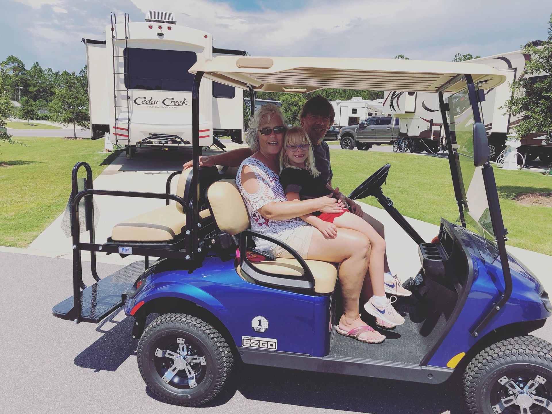 A blue golf cart holding three people sits in a sunny RV park.