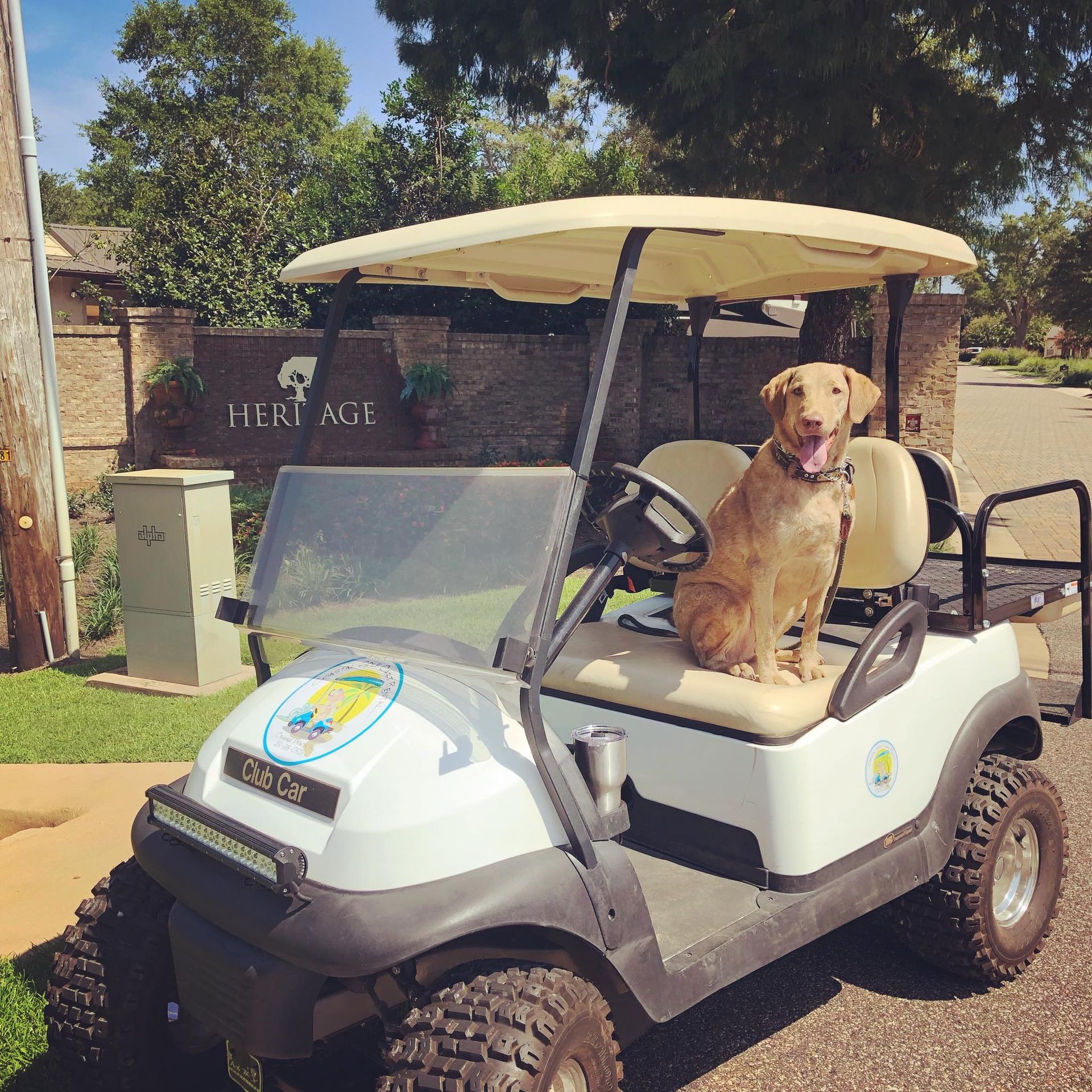 A light brown dog sits on the driver's seat of a white golf cart parked in front of a Heritage community brick sign.