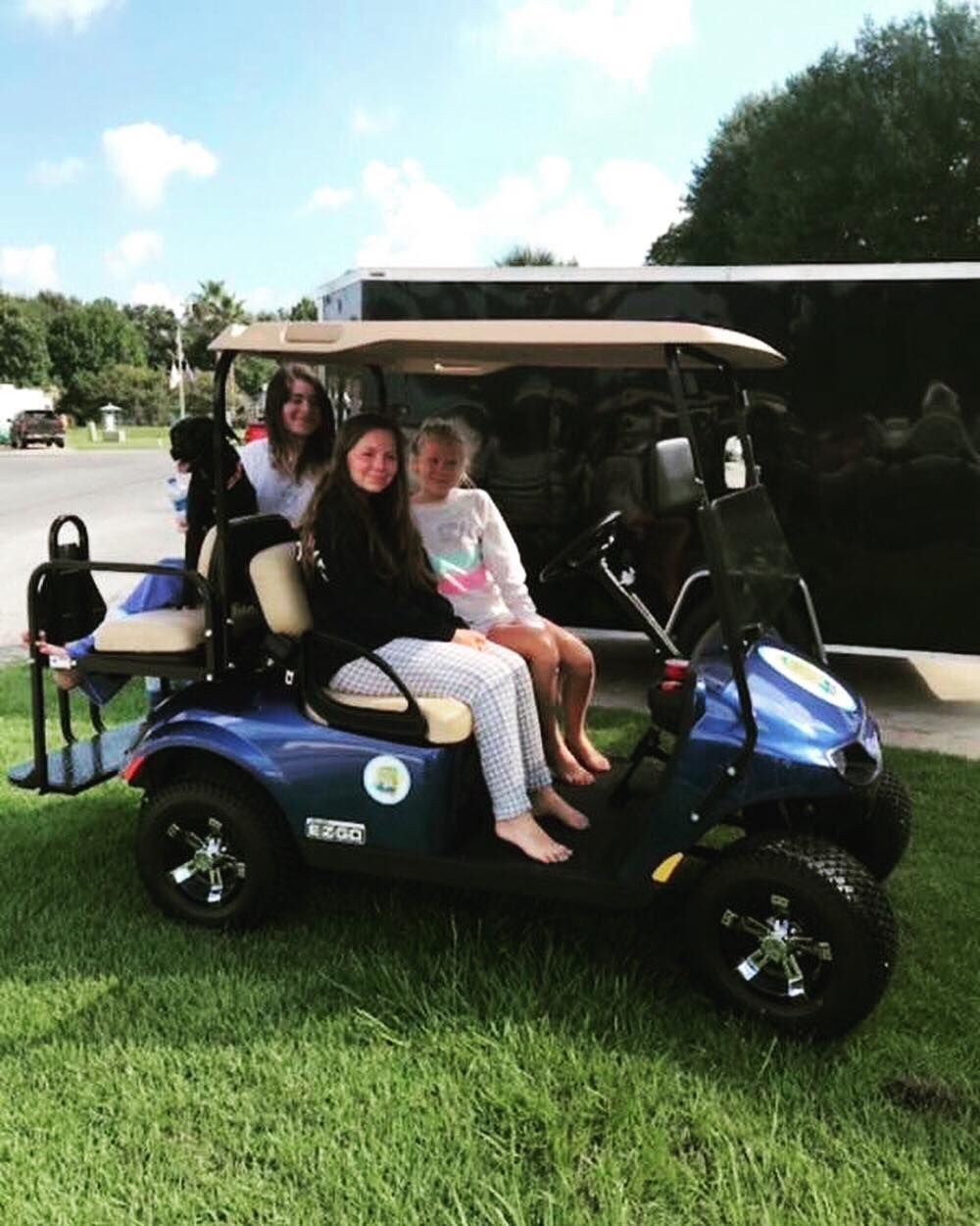 Three people sit in a blue golf cart on grass next to a black trailer under a sunny sky.
