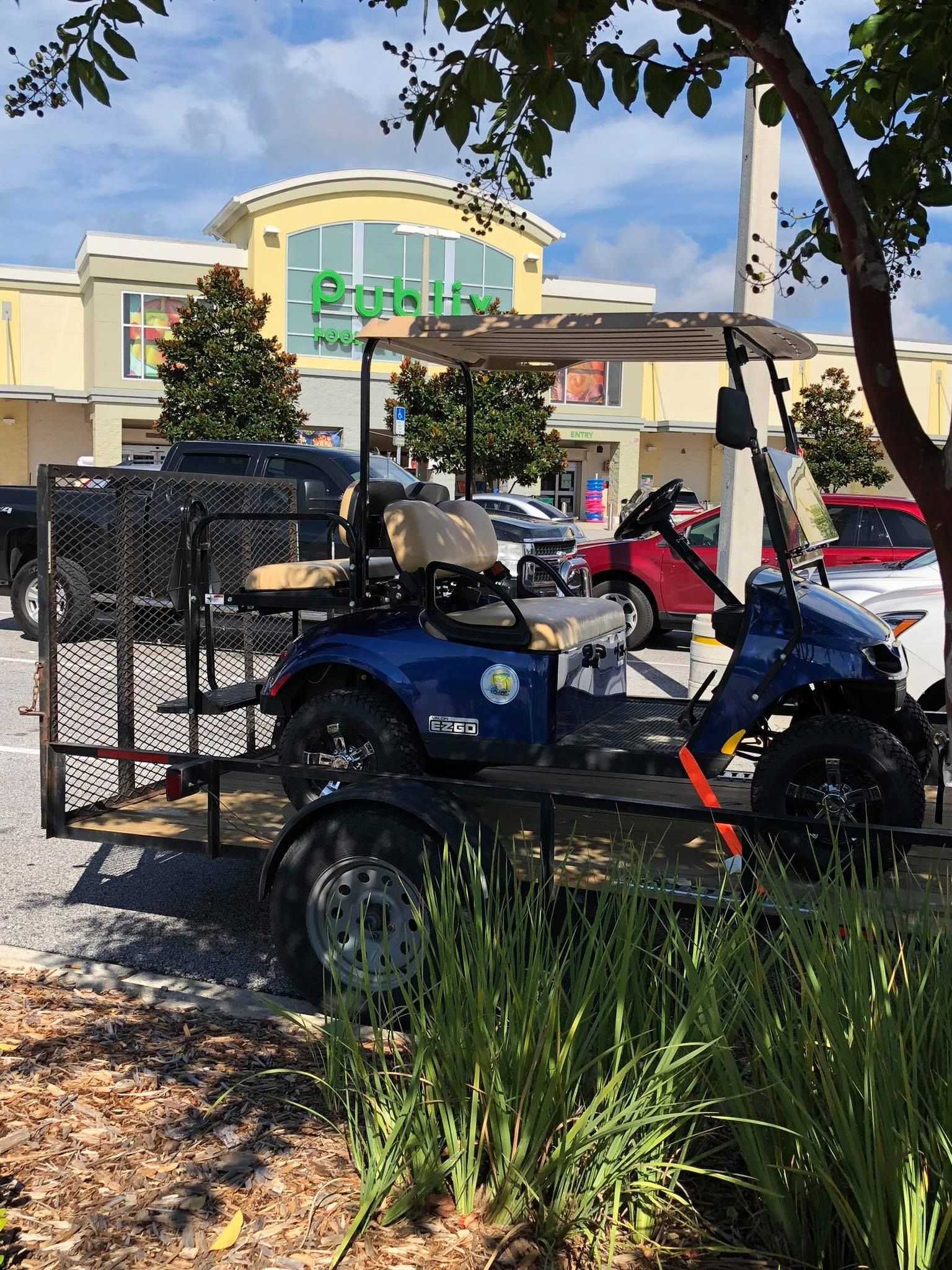 A blue golf cart loaded on a black metal utility trailer, parked in front of a Publix grocery store under a sunny sky.