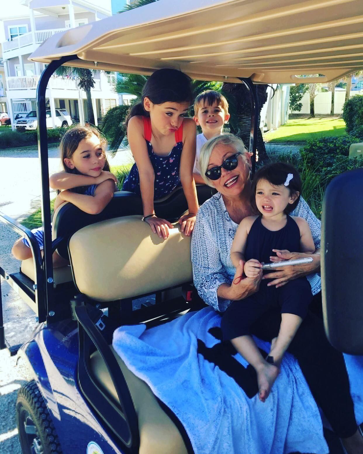 A group of people sitting in a golf cart, including a smiling adult holding a young child, outdoors on a sunny day.