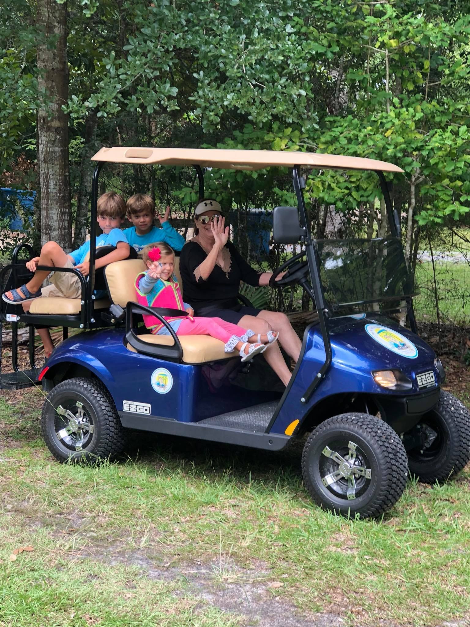 Four people riding in a blue golf cart outdoors; some are waving at the camera.