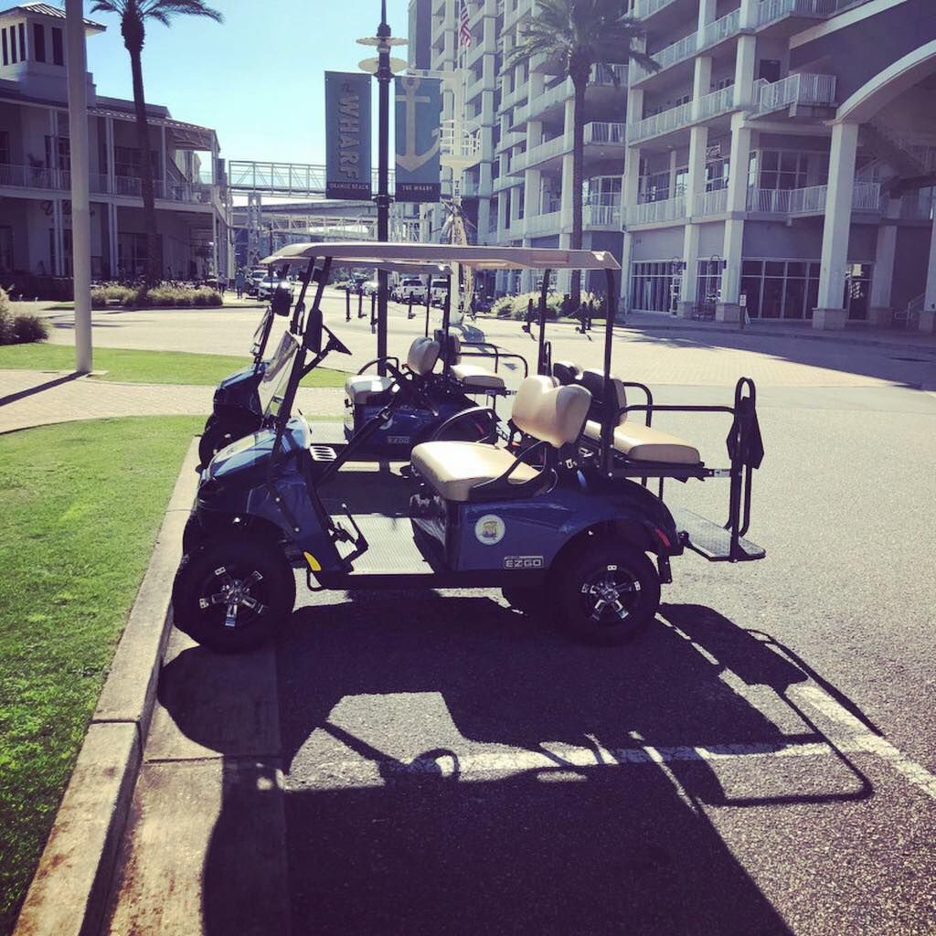 Two blue four-seater golf carts parked on the curb of a sunny, paved plaza near a multi-story building.
