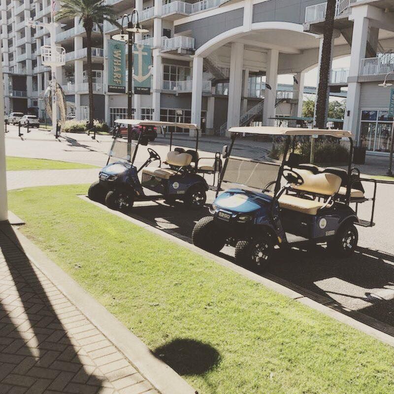 Two dark blue golf carts parked on a paved path next to a grassy lawn in front of a multi-story building at The Wharf.