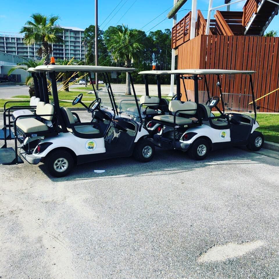 Three white golf carts are parked in a line on an asphalt lot near a wooden structure and palm trees.