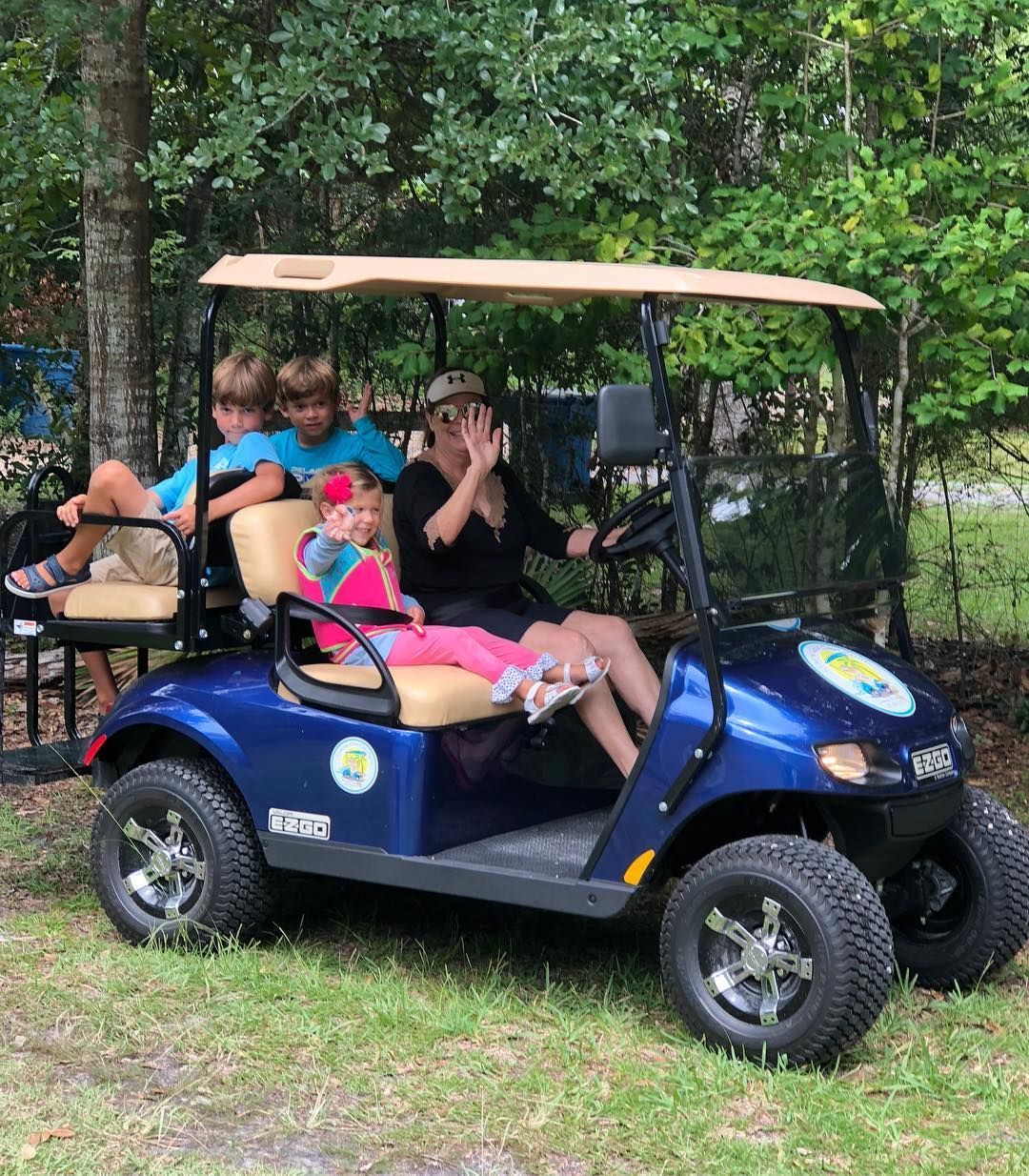 A group of people sits in a blue golf cart parked on grass near trees, with one person waving.