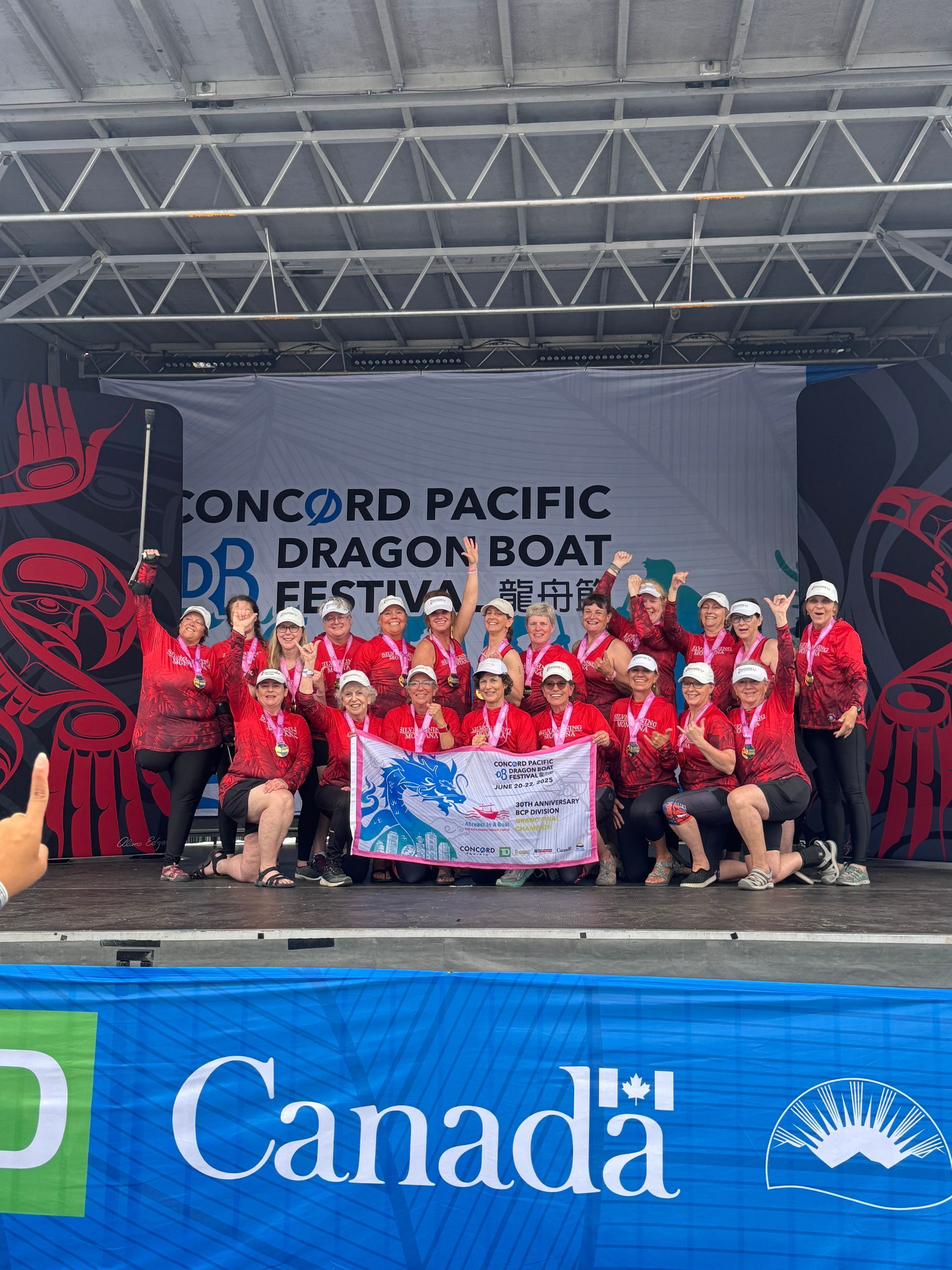 Dragon boat team celebrates on stage at Concord Pacific Festival, red and white outfits, banner, Canada sign.