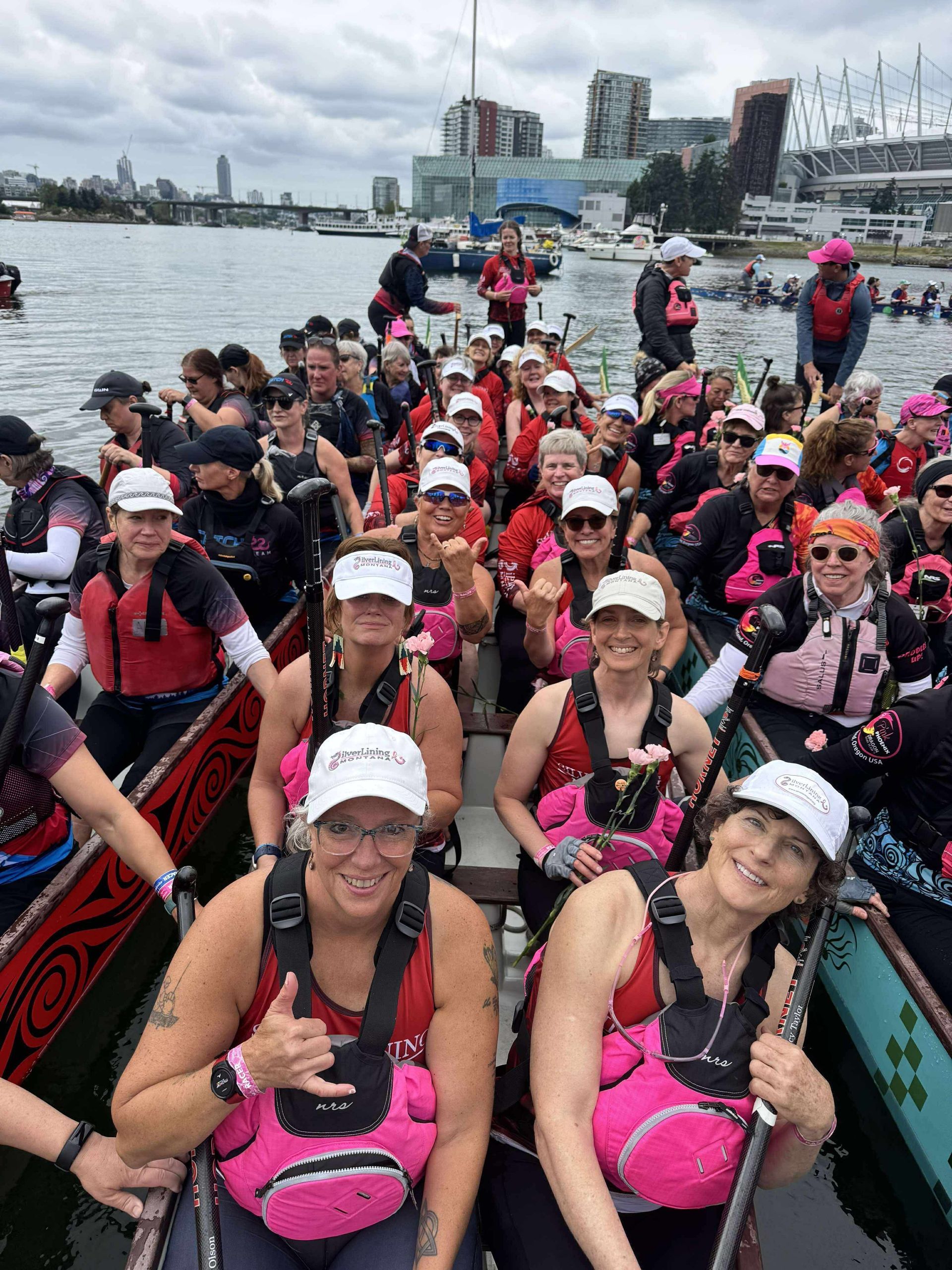 Dragon boat team, women in pink life vests and hats, smiling, in a boat on water, buildings in the background.