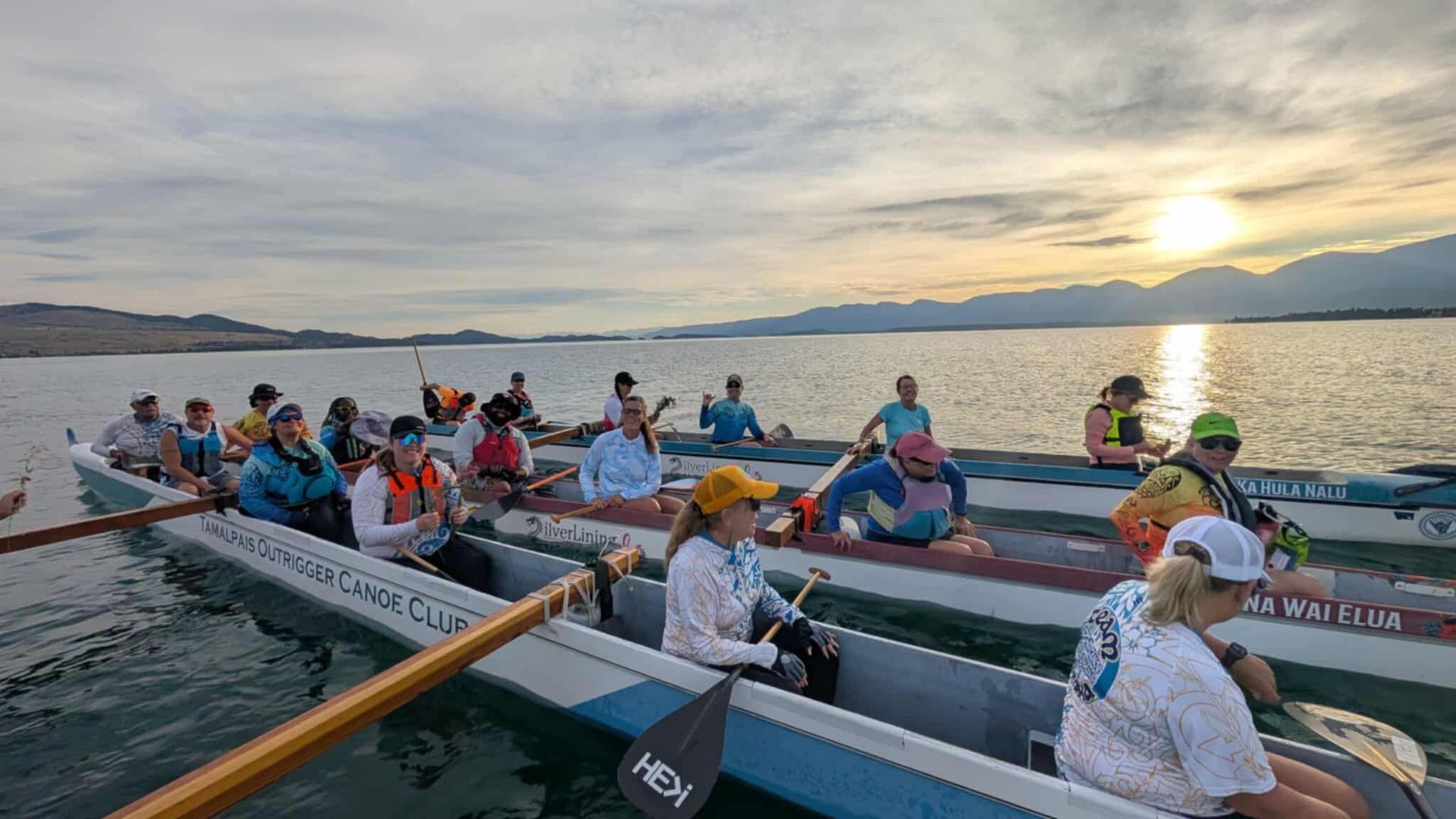 Canoe race on lake at sunset. People paddling in long canoes. Mountains in the background.