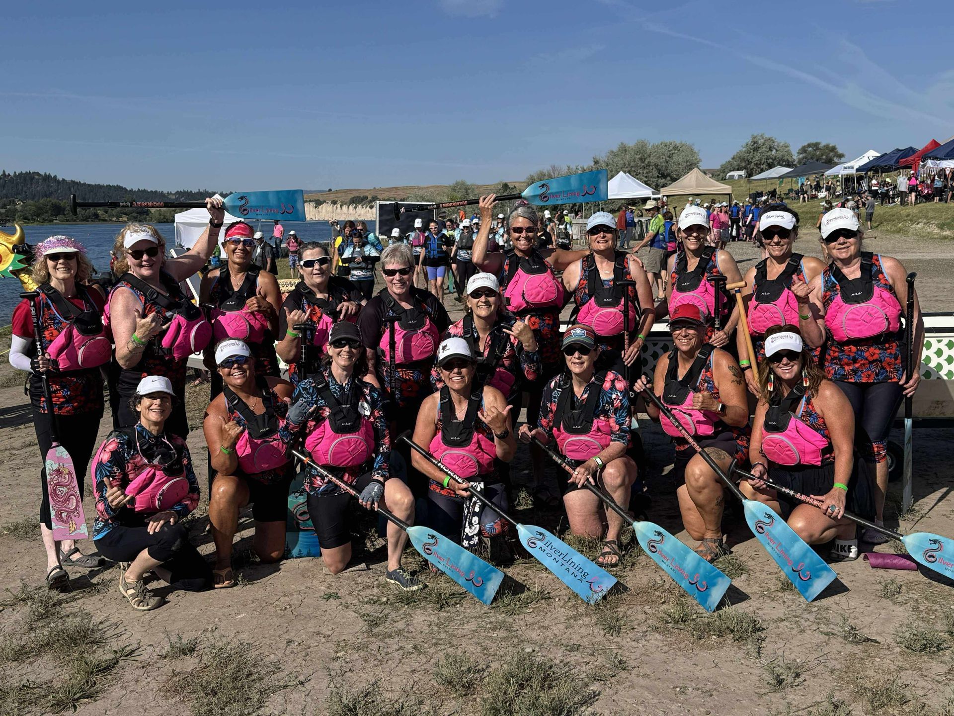 Group of women in pink life vests and hats pose with paddles by a lake.