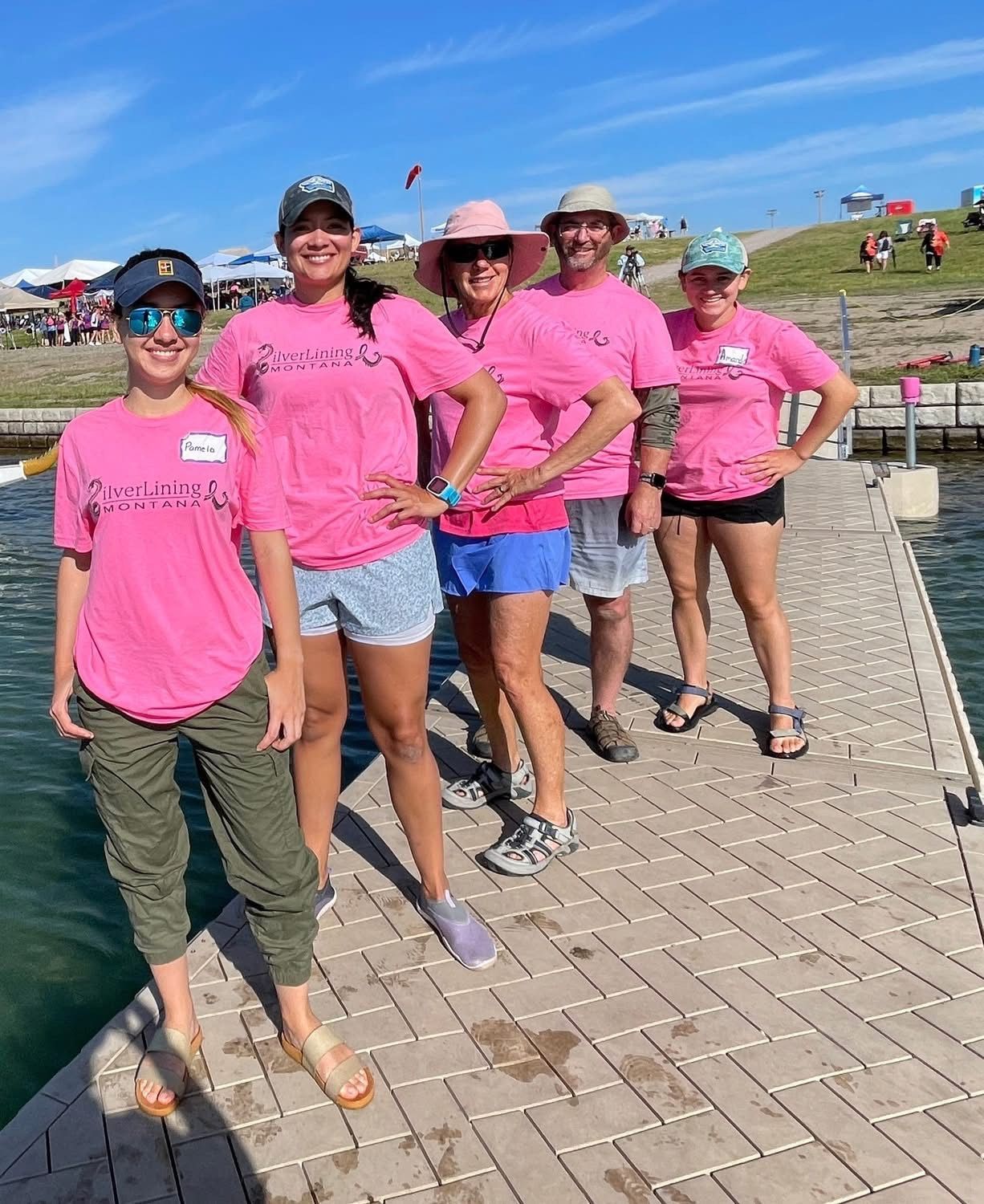 Group of five people wearing pink shirts on a wooden dock, smiling at the camera under a blue sky.