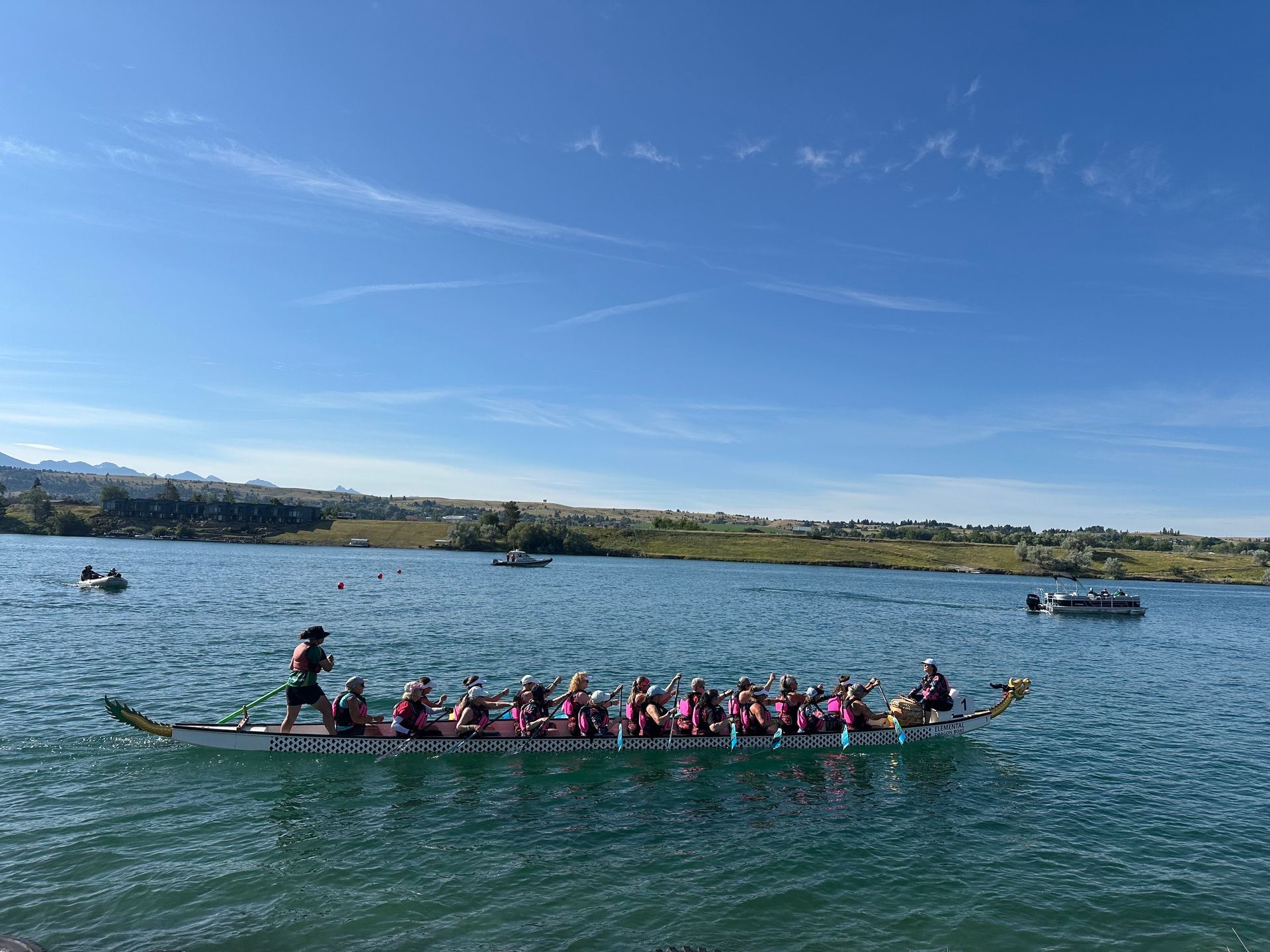 Dragon boat on a lake with paddlers, a drummer, and a steersperson under a blue sky.