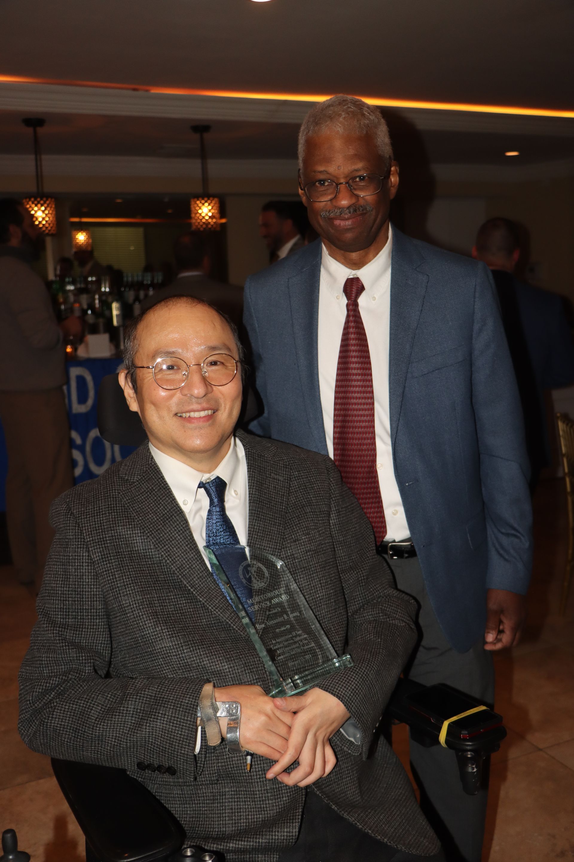 A Man In A Suit And Tie Is Standing Next To A Man In A Wheelchair Holding A Trophy — Staten Island, NY — RCBA