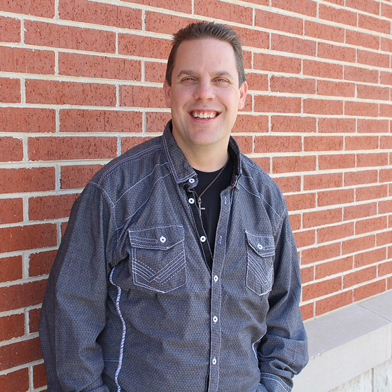 A man in a denim shirt is smiling in front of a brick wall