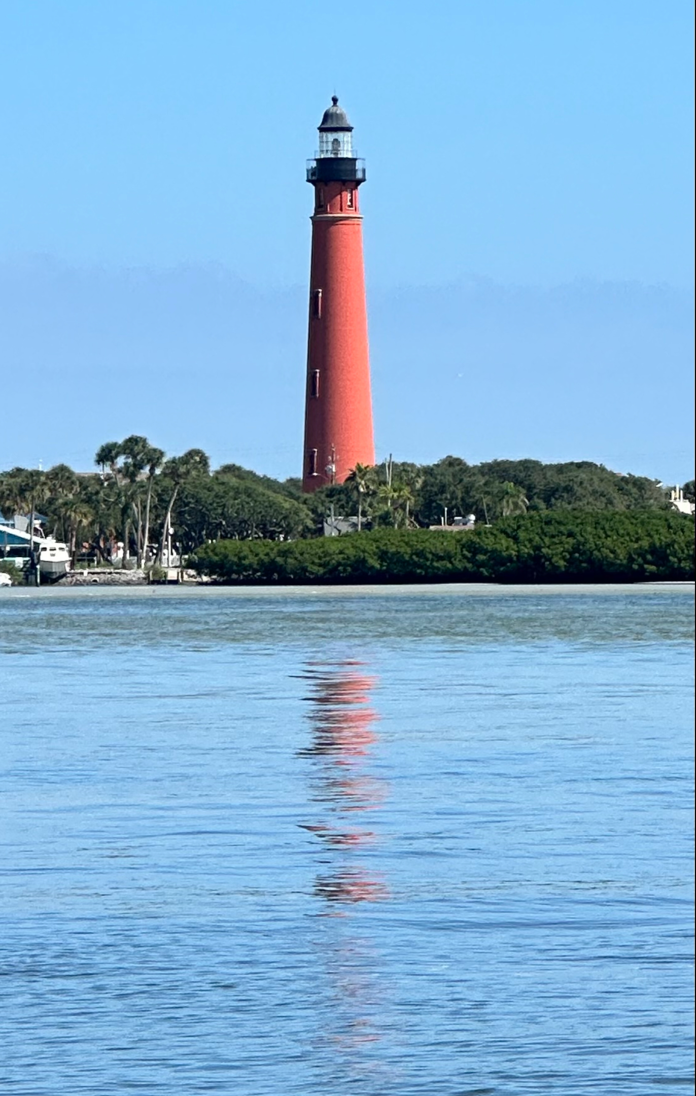 Red lighthouse on a green treed island, with the reflection in the blue water.