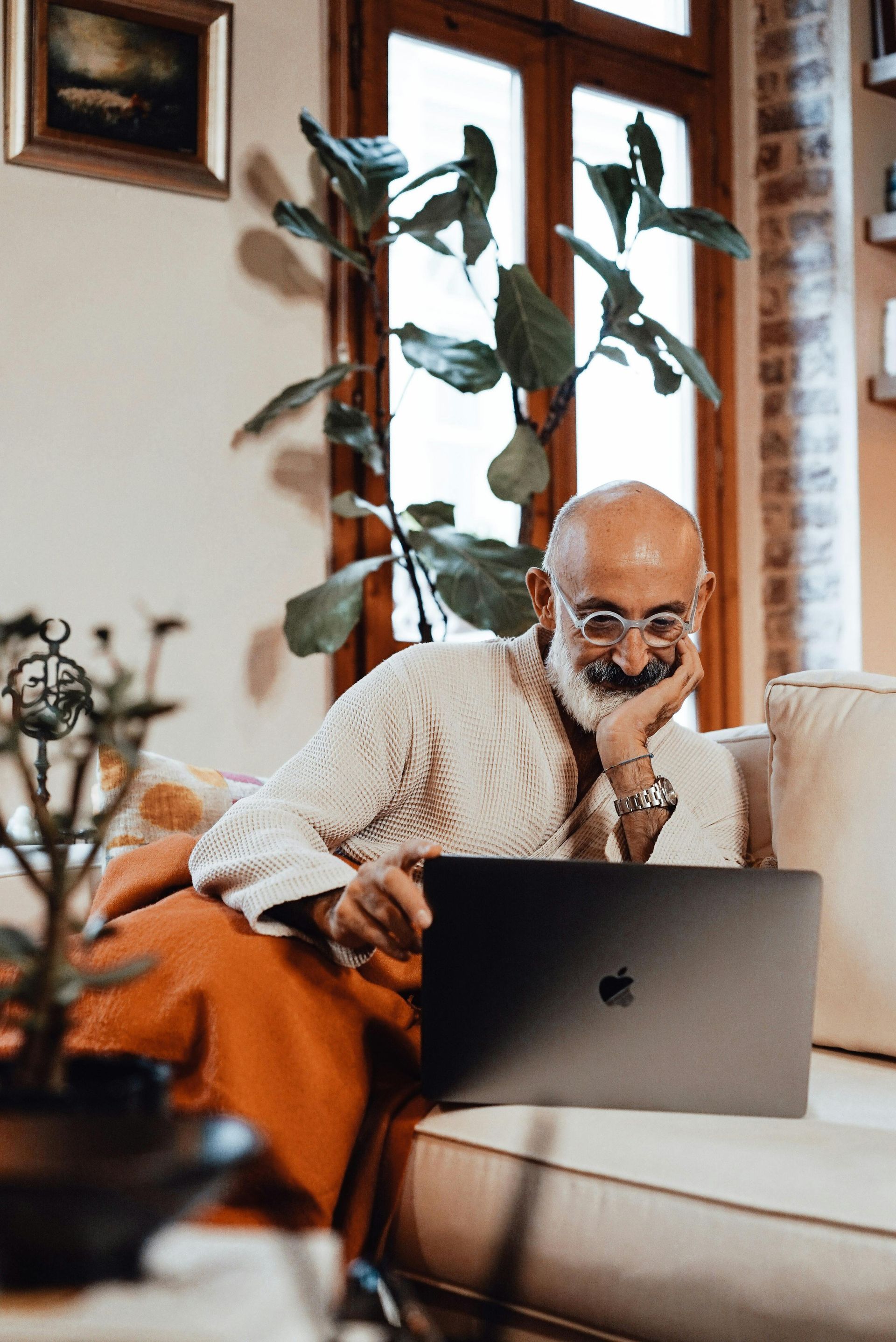 A person with a beard and glasses sits on a sofa, smiling while looking at an open laptop in a sunlit living room.