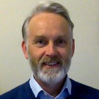 Man with a graying beard smiles at the camera, wearing a blue sweater and collared shirt.