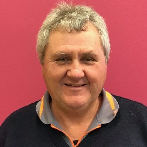 Man with gray hair, smiling, wearing a navy sweater with an orange-trimmed collar, pink background.