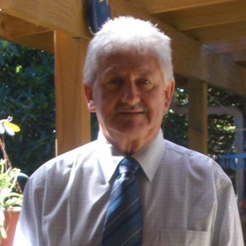 Man wearing a tie and collared shirt stands on a porch; sunlight on face, neutral expression.
