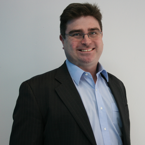 Man in glasses, blue shirt, and black suit jacket smiles at the camera against a white background.