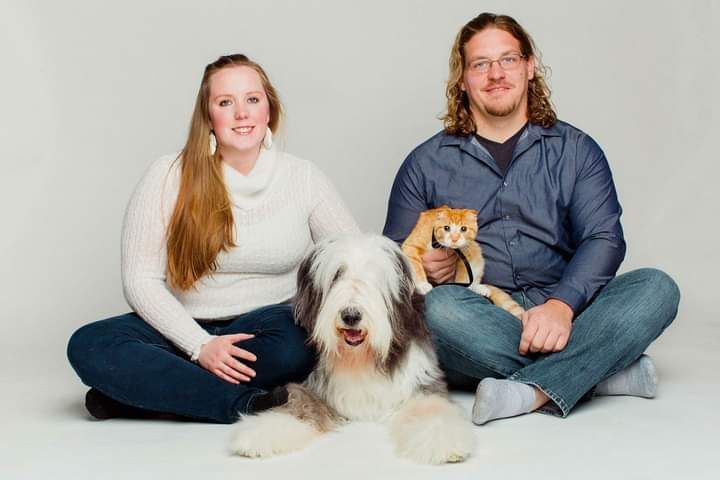A woman and a man sit on the floor with a large, shaggy dog and an orange cat in a studio setting.