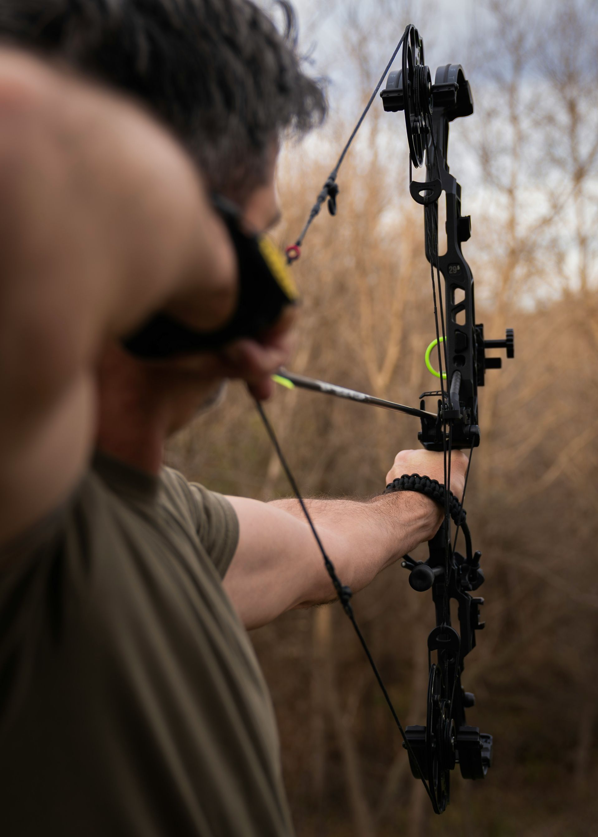 Person drawing a black compound bow, aiming an arrow. Outdoors with trees in the background.