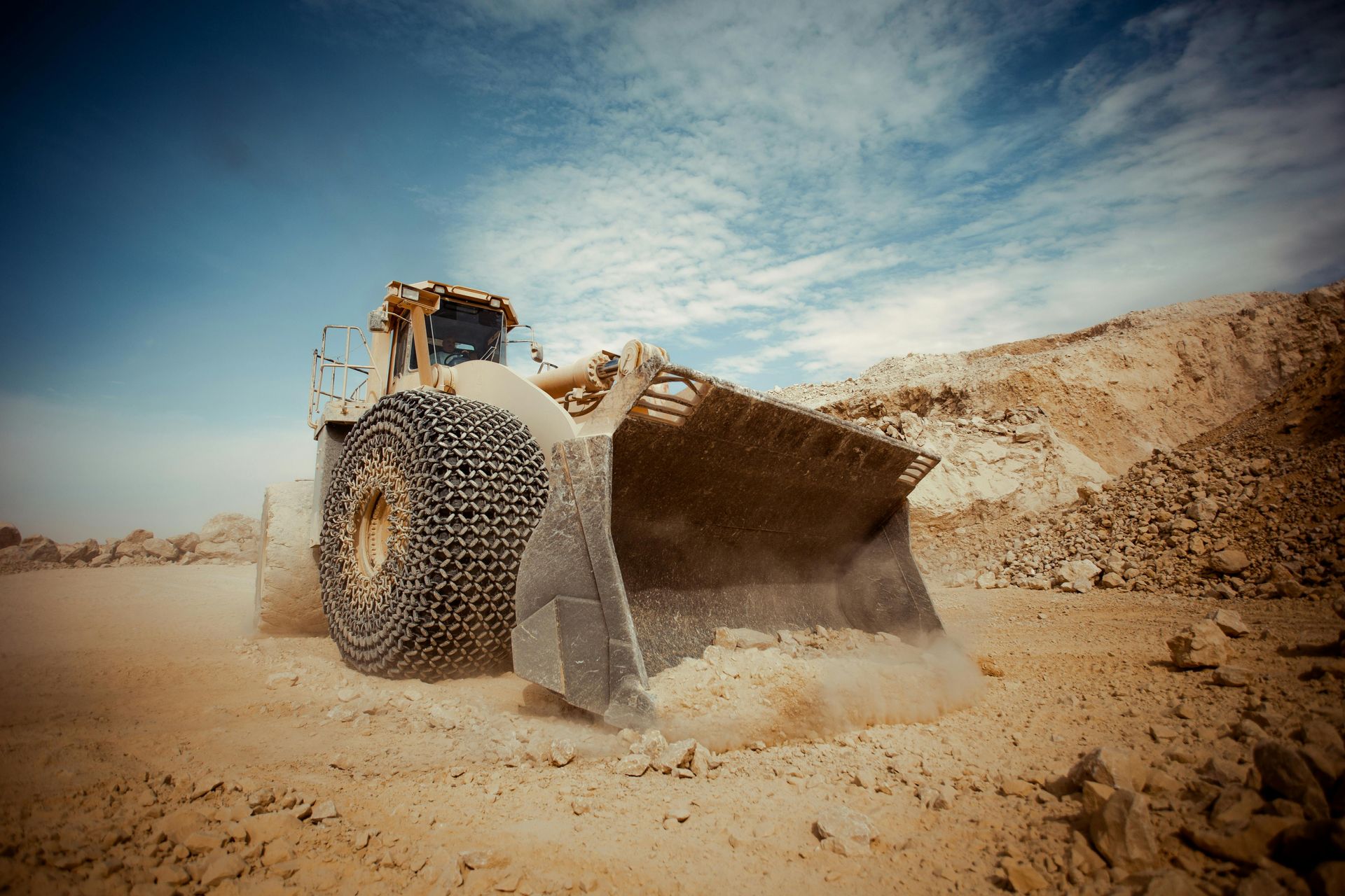 A large front-end loader scoops earth at a construction site under a cloudy blue sky.