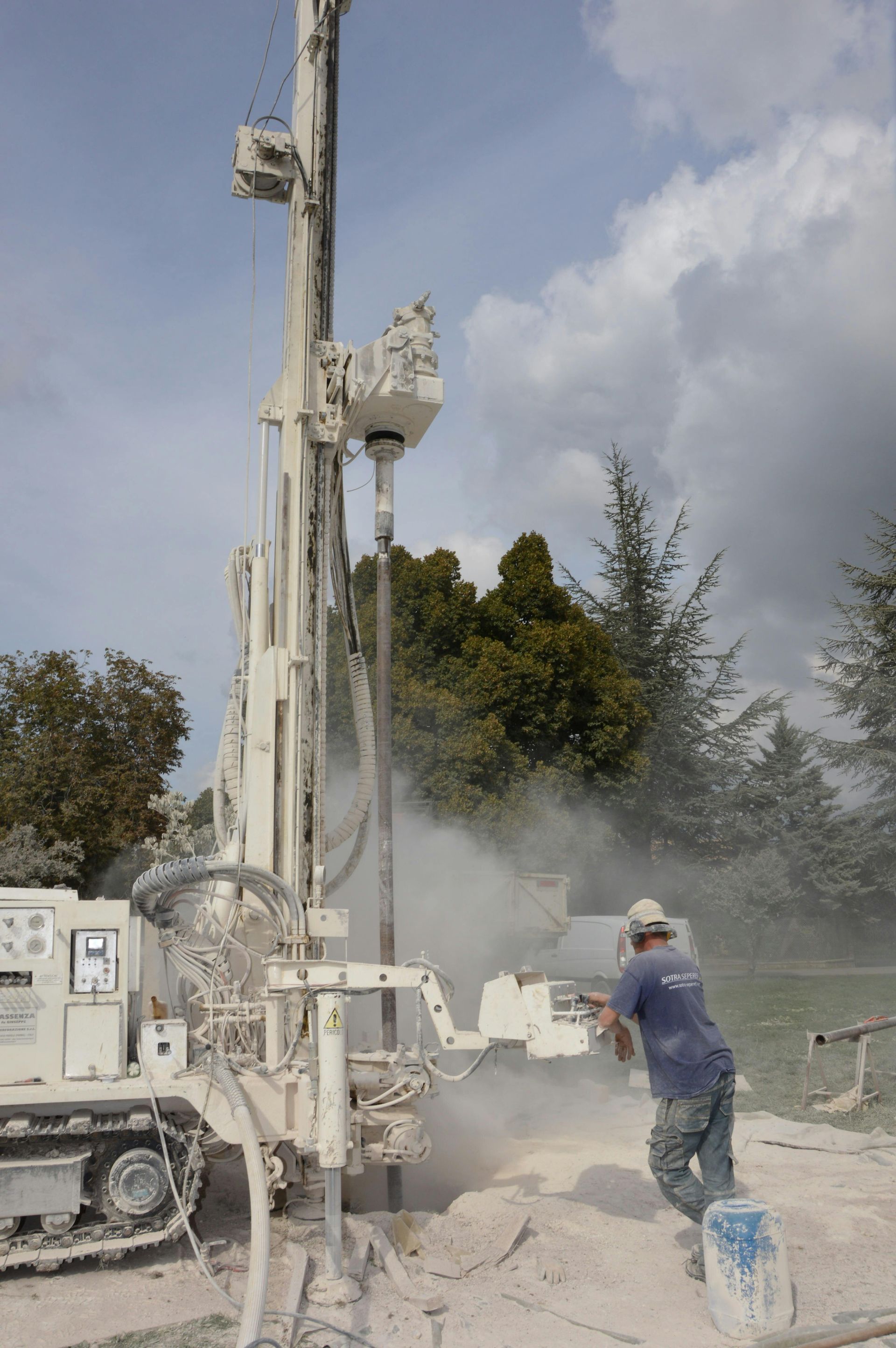 A worker operating a drilling rig, creating dust. The machine is white, the setting is outdoors.