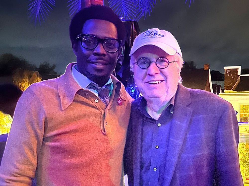 Two men are posing for a picture together . one of the men is wearing glasses and a hat.