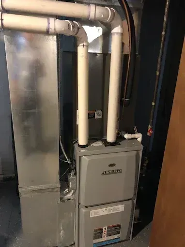 Furnace unit with two white pipes, a silver flue and ductwork, in a basement.