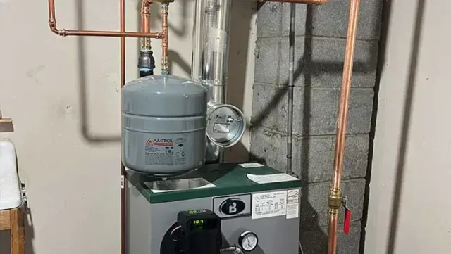 Water heater with copper pipes in a basement. Grey tank and black control panel, with a red valve.