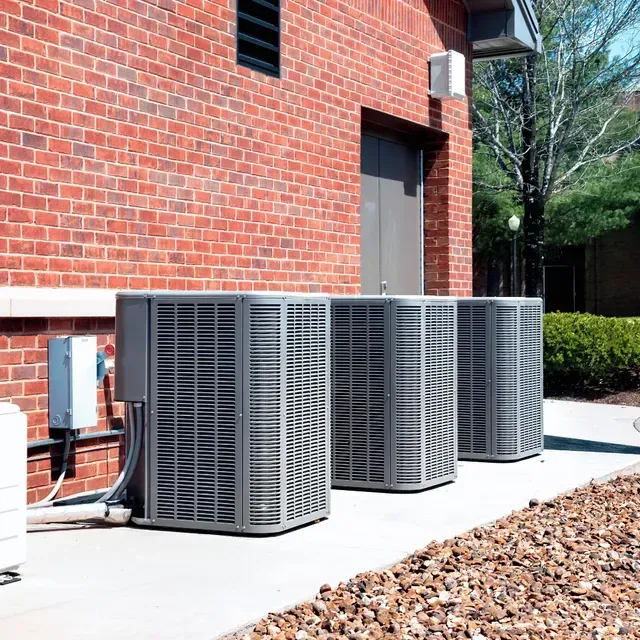 Three air conditioning units outside a brick building, on a concrete pad.