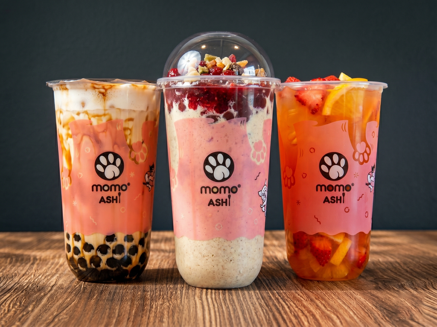 Three Momo Ashi bubble tea drinks in plastic cups with fruit, tapioca pearls, and toppings on a wooden table.