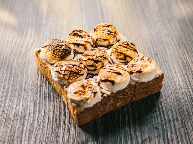 A piece of toasted bread topped with nine toasted marshmallows drizzled with chocolate, set on a wood-grain surface.