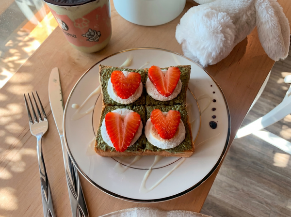 A piece of toast topped with matcha spread, cream, and heart-shaped strawberry slices, served on a plate with cutlery.