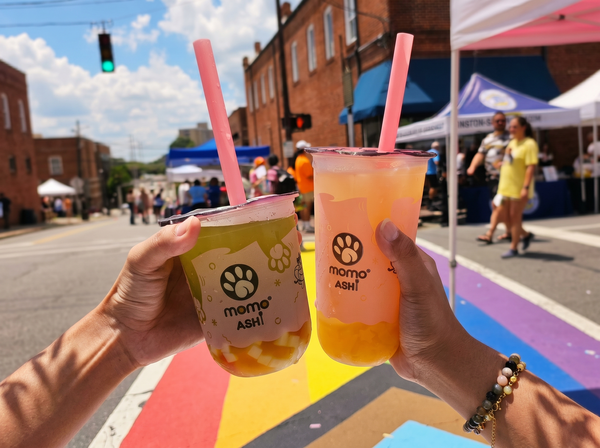 Two hands hold bubble tea cups with pink straws against a sunny, outdoor street scene with a painted rainbow crosswalk.