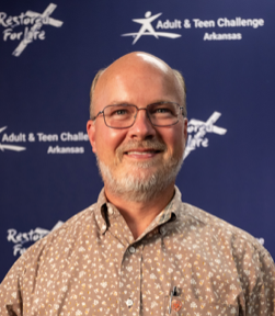 A man wearing glasses stands in front of a sign that says adult & teen challenge