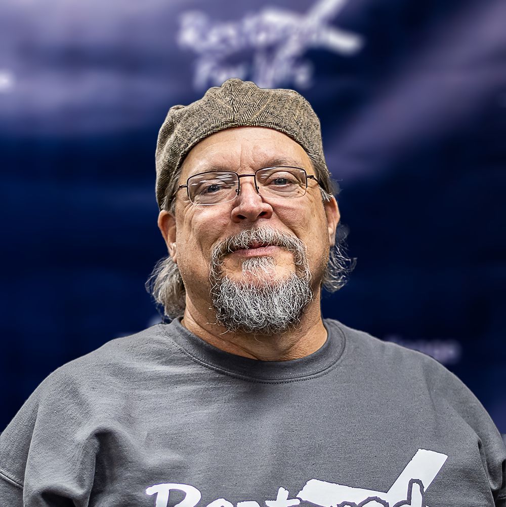 A man with a beard and glasses is wearing a hat and a gray shirt.