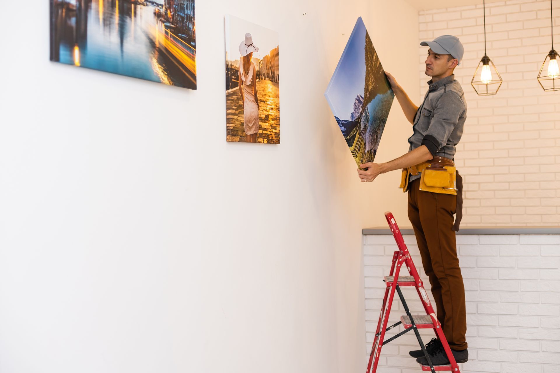 Man on ladder hangs artwork on white wall, wearing tool belt and cap.