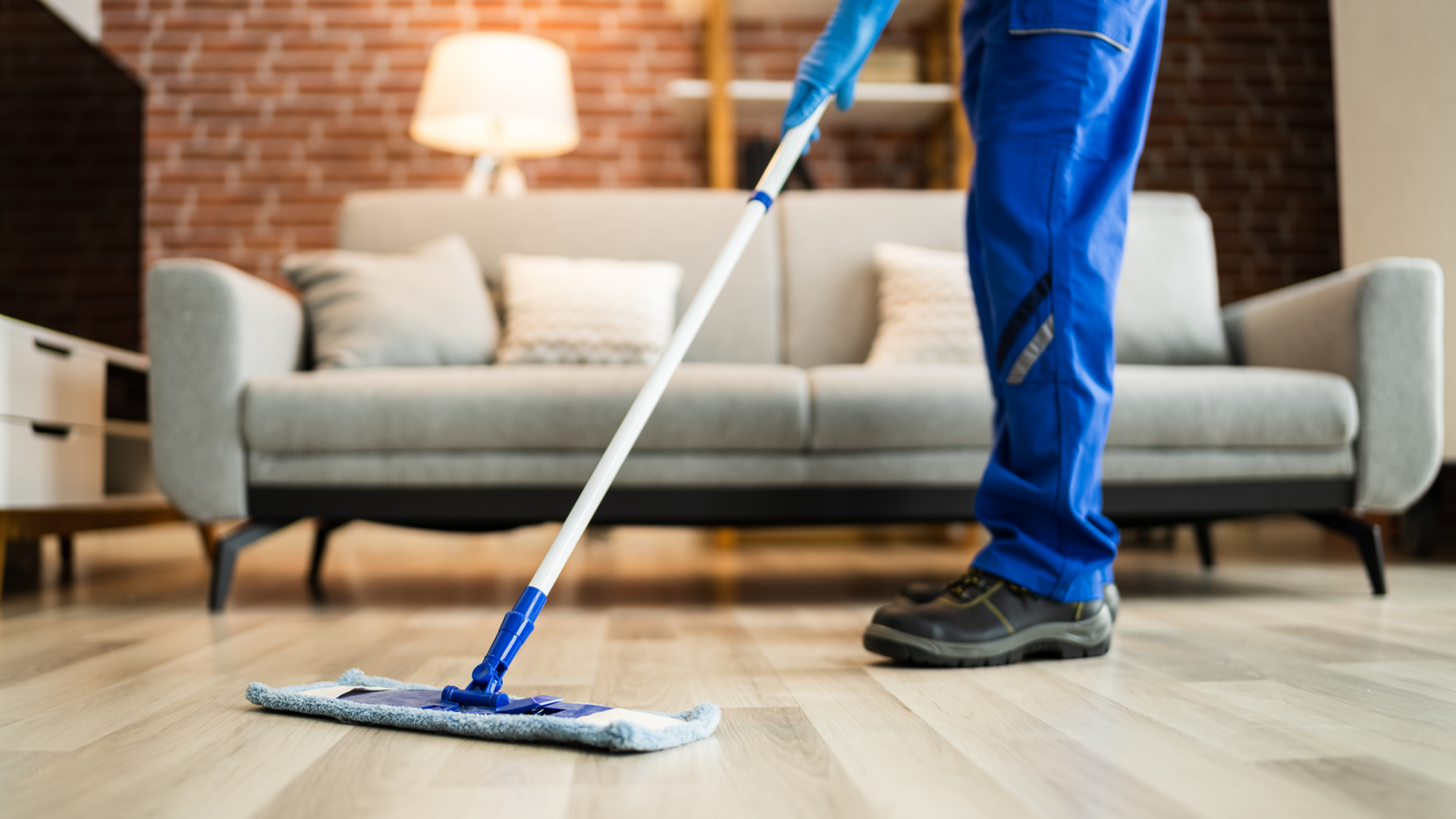 Person mopping a hardwood floor in a living room with a couch and lamp nearby