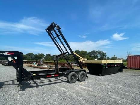 A dumpster is being pulled by a trailer in a gravel lot.