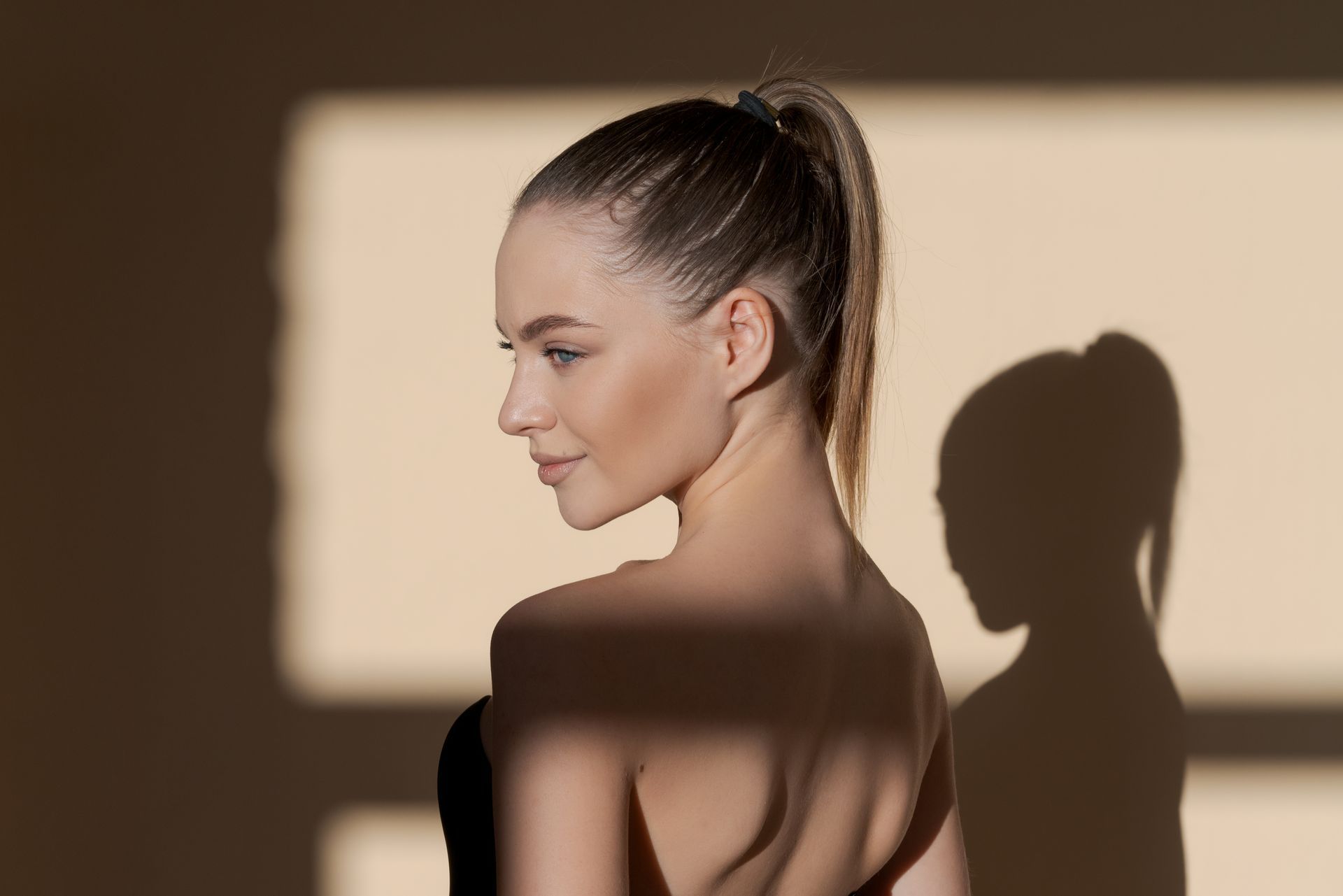 A woman with a ponytail is standing in front of a wall with a shadow on her back.