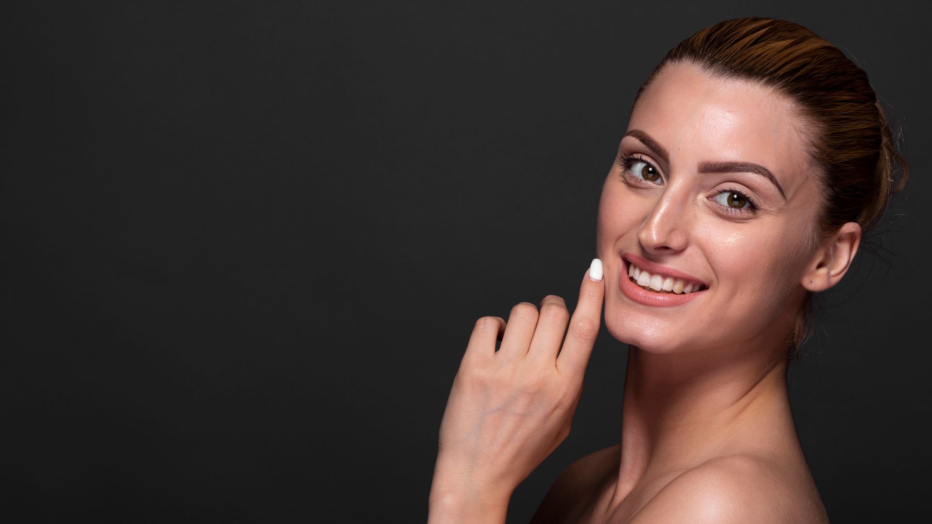 Woman with fair skin, smiling and touching her cheek against a dark gray background.