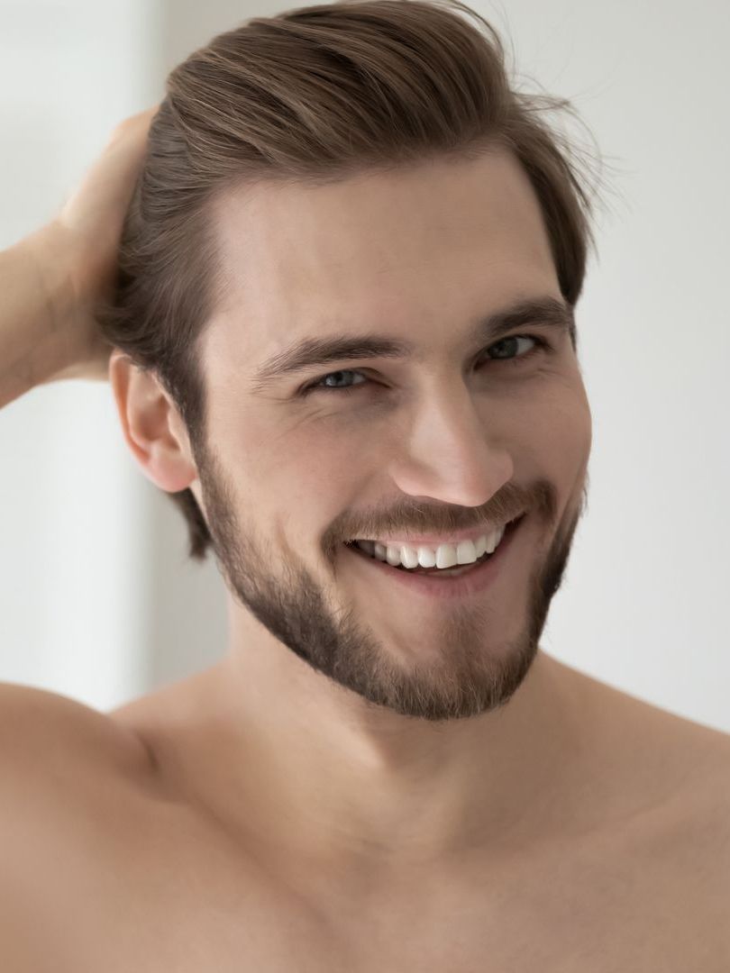 A shirtless man with a beard is smiling and touching his hair.