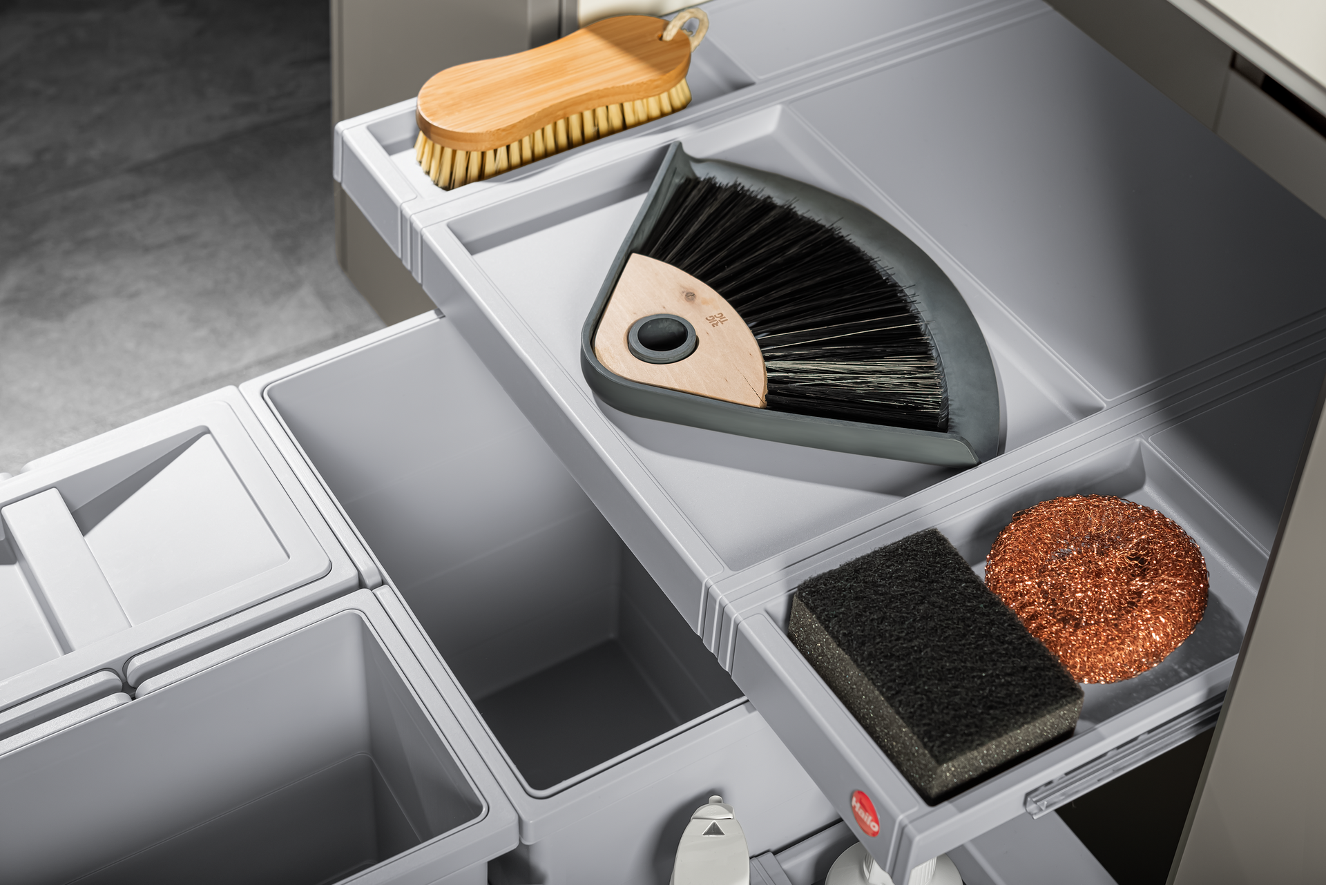Gray pull-out kitchen cabinet drawer with cleaning supplies: scrub brush, dustpan, sponge, and scrubber.